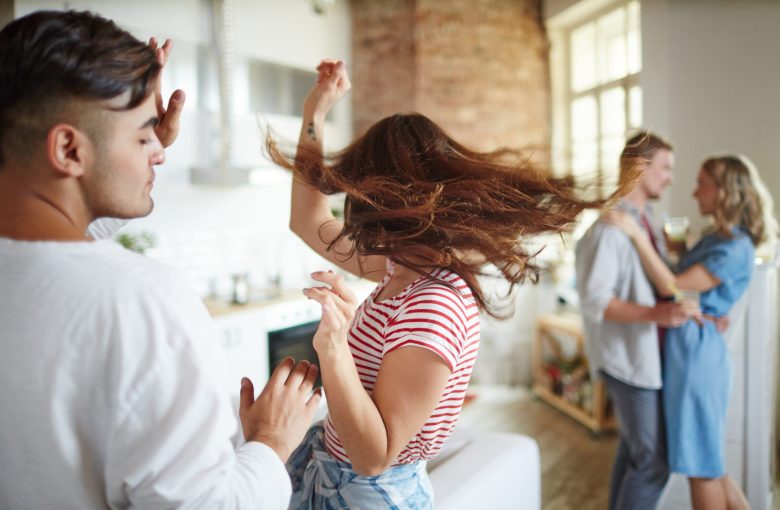 Ecstatic girl shaking her long hair while dancing at home party with her boyfriend near by and amorous couple on background